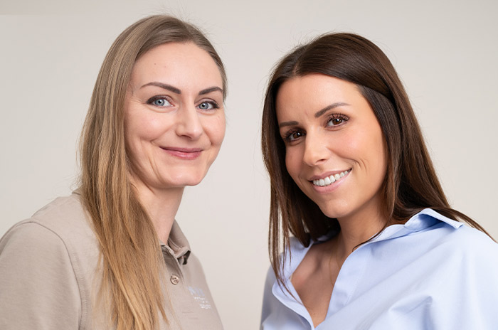 Zwei lächelnde nebeneinanderstehende Frauen vor beigem Hintergrund